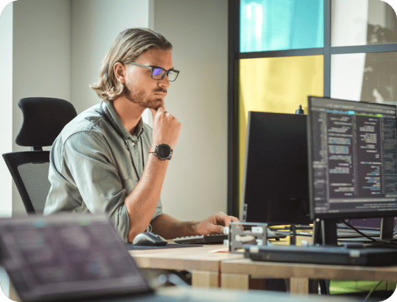 Ein Mann mit Brille sitzt an einem Schreibtisch und arbeitet an zwei nebeneinander stehenden Computern.